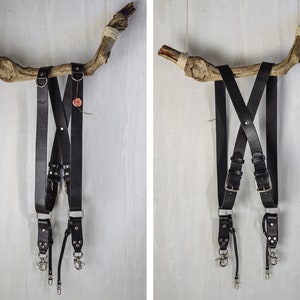 Puede incluir: Dos correas de cámara de cuero negro con herrajes plateados colgando de un trozo de madera a la deriva. Las correas están cruzadas en forma de X y tienen hebillas ajustables.