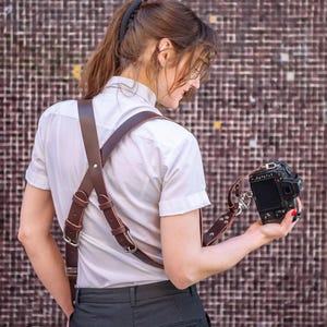 Op de afbeelding: Een vrouw draagt een wit shirt en een zwarte broek met een bruine leren cameraband. Ze houdt een zwarte camera in haar rechterhand.