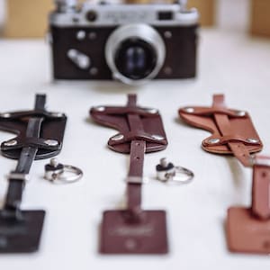 Può includere: Tre cinturini per fotocamera in pelle nera, marrone e marrone chiaro, ciascuno con un portachiavi argentato. Una fotocamera vintage è sullo sfondo. I cinturini hanno un'etichetta rettangolare in basso. Il logo Miller Things si trova in basso a destra.