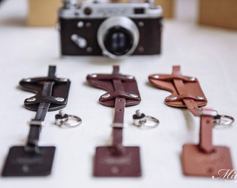 May include: Three leather camera straps in black, brown, and tan, each with a silver-toned key ring. A vintage camera is in the background. The straps have a rectangular tag at the bottom. The Miller Things logo is in the bottom right corner.