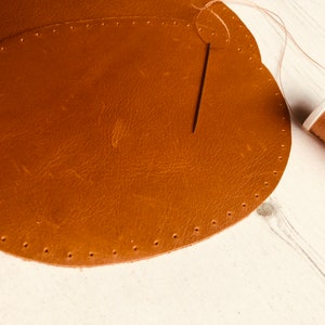 May include: Two brown leather patches with holes around the edges, a needle and thread are being used to sew the patches together.