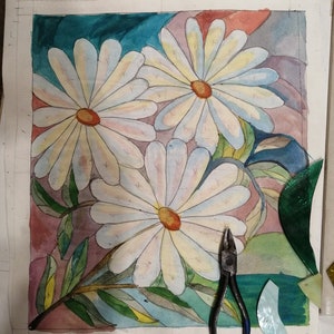 May include: A watercolor painting of three white daisies with yellow centers on a background of teal, pink, and gray. The painting is on a white sheet of paper with a pair of pliers and pieces of stained glass on the table.