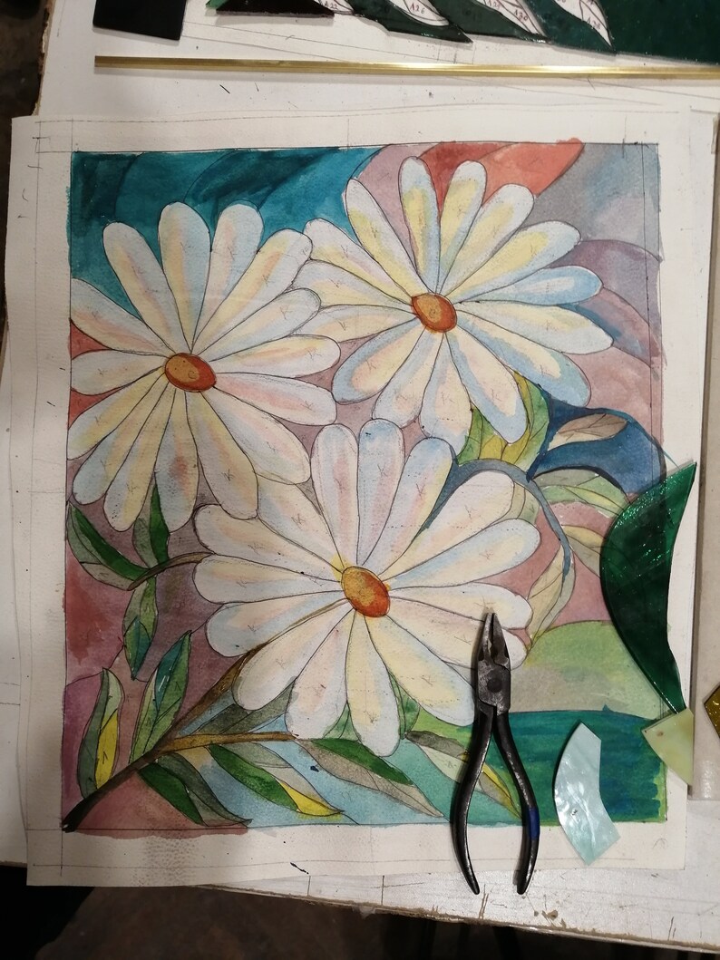 May include: A watercolor painting of three white daisies with yellow centers on a background of teal, pink, and gray. The painting is on a white sheet of paper with a pair of pliers and pieces of stained glass on the table.