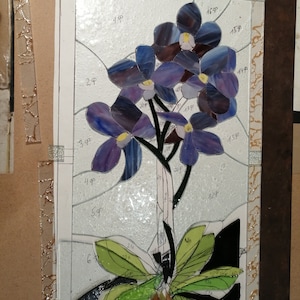 May include: A stained glass artwork depicting a purple orchid with green leaves. The artwork is framed and has numbers written on it.