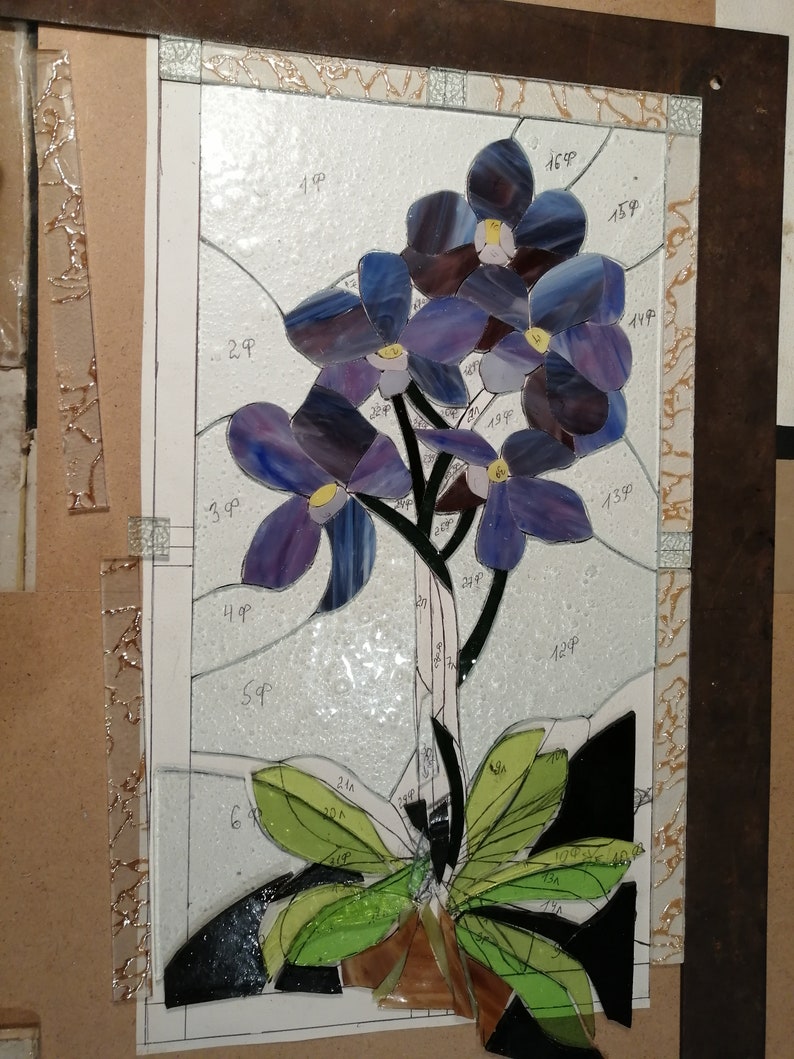 May include: A stained glass artwork depicting a purple orchid with green leaves. The artwork is framed and has numbers written on it.