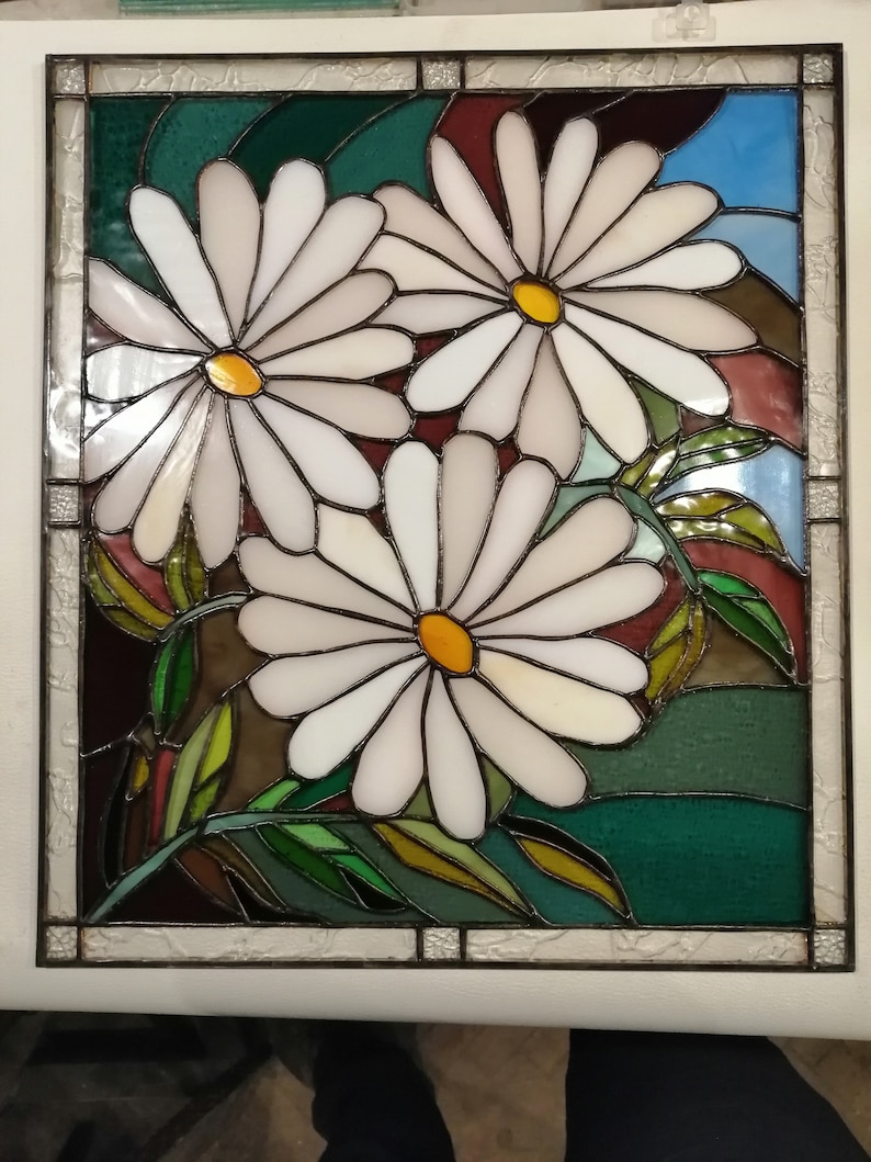 May include: A stained glass window featuring three white daisies with yellow centers. The daisies are set against a background of green, brown, and blue glass.