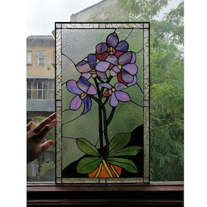 May include: A stained glass window panel depicting a purple orchid plant in a pot. The orchid has several flowers with pink and purple shades. The panel is framed in gold.