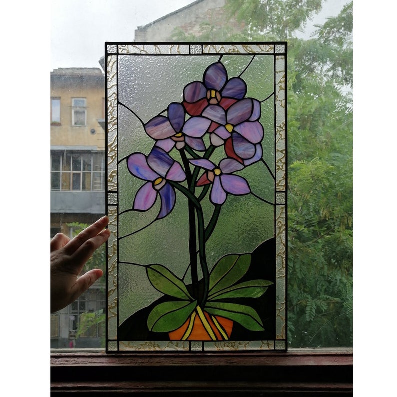 May include: A stained glass window panel depicting a purple orchid plant in a pot. The orchid has several flowers with pink and purple shades. The panel is framed in gold.