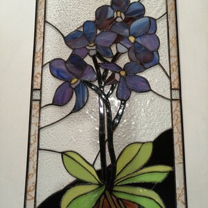 May include: A stained glass window featuring a bunch of purple orchids with green leaves in a brown pot. The window is framed in black and has a textured glass background.