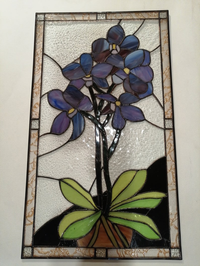 May include: A stained glass window featuring a bunch of purple orchids with green leaves in a brown pot. The window is framed in black and has a textured glass background.