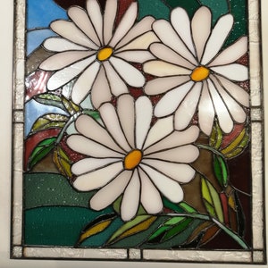 May include: A stained glass window featuring three white daisies with yellow centers, surrounded by green leaves and a blue sky background.