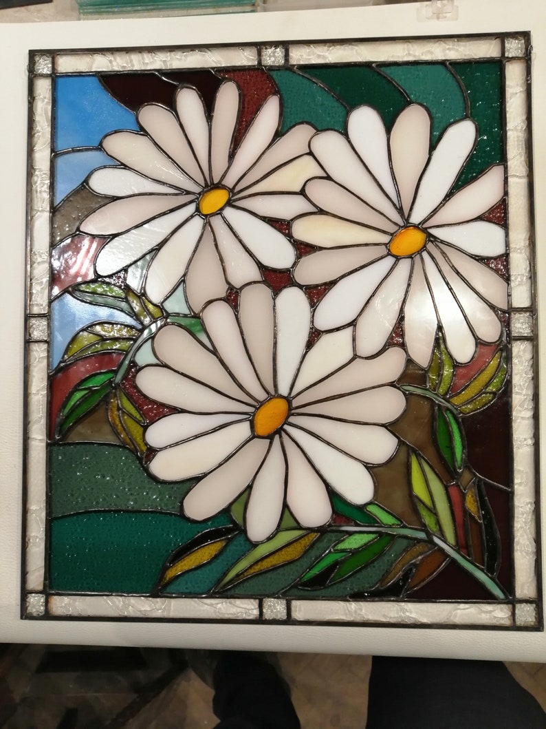 May include: A stained glass window featuring three white daisies with yellow centers, surrounded by green leaves and a blue sky background.