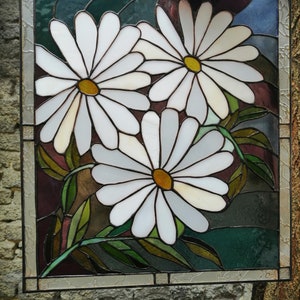 May include: A stained glass window panel featuring three white daisies with yellow centers, surrounded by green leaves on a dark background.