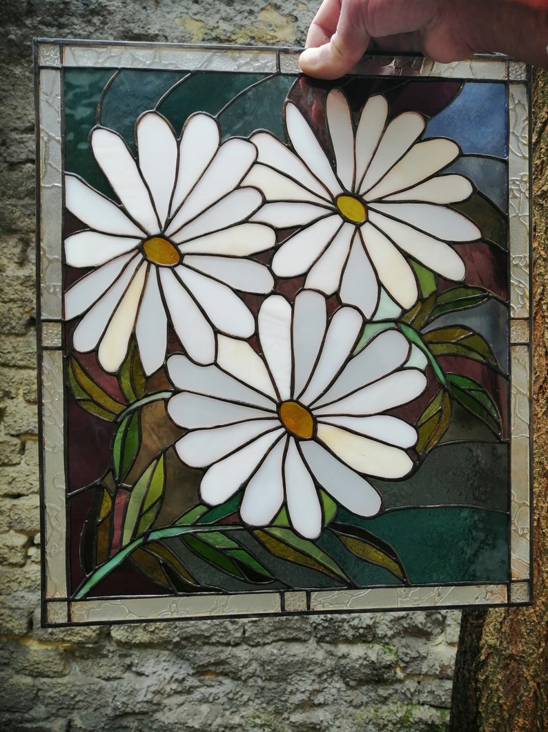 May include: A stained glass window panel featuring three white daisies with yellow centers, surrounded by green leaves on a dark background.