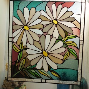 May include: A stained glass window featuring three white daisies with yellow centers, set against a background of teal, pink, and brown. The daisies are arranged in a cluster, with their stems and leaves visible.