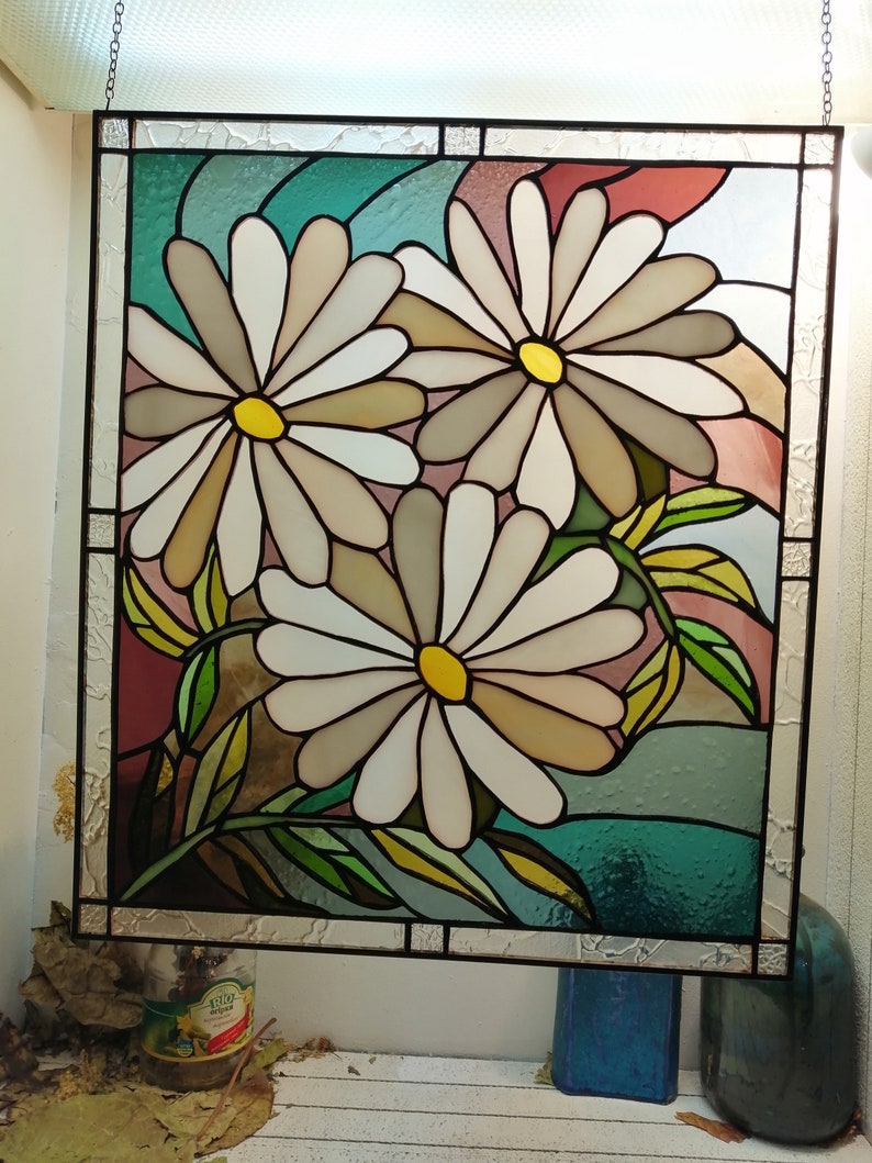 May include: A stained glass window featuring three white daisies with yellow centers, set against a background of teal, pink, and brown. The daisies are arranged in a cluster, with their stems and leaves visible.