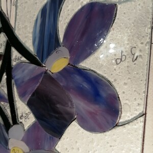 May include: Close-up of a stained glass mosaic art piece featuring purple and blue flowers with yellow centres. The piece is set against a white background.