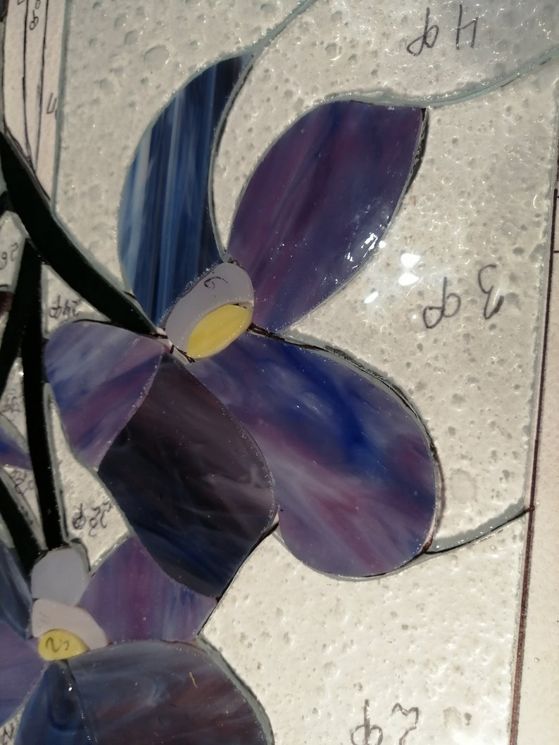May include: Close-up of a stained glass mosaic art piece featuring purple and blue flowers with yellow centres. The piece is set against a white background.