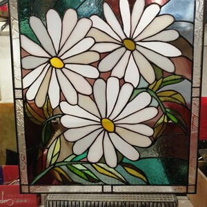 May include: A stained glass window depicting three white daisies with yellow centers. The daisies are surrounded by green leaves and a background of blue, green, and red glass.