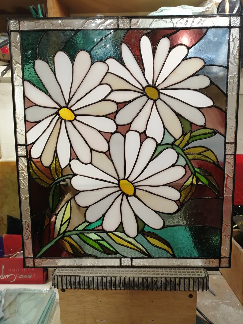 May include: A stained glass window depicting three white daisies with yellow centers. The daisies are surrounded by green leaves and a background of blue, green, and red glass.