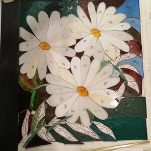 May include: A stained glass mosaic art piece depicting three white daisies with yellow centers. The daisies are surrounded by green leaves and a blue background. The glass pieces are numbered.