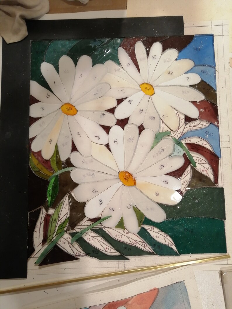 May include: A stained glass mosaic art piece depicting three white daisies with yellow centers. The daisies are surrounded by green leaves and a blue background. The glass pieces are numbered.