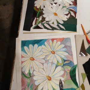 May include: A close-up of a watercolor painting of white daisies with green leaves on a pink and blue background. A pair of pliers is on the right side of the painting. There are also pieces of glass in the background.