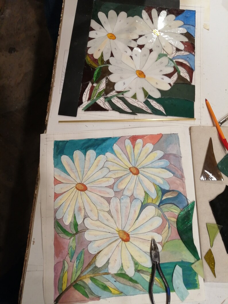 May include: A close-up of a watercolor painting of white daisies with green leaves on a pink and blue background. A pair of pliers is on the right side of the painting. There are also pieces of glass in the background.