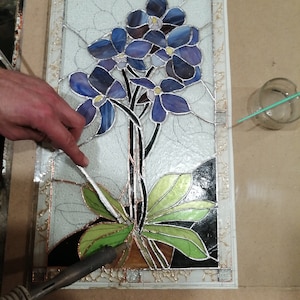 May include: A stained glass panel depicting a cluster of blue flowers with green leaves. The panel is being assembled with a soldering iron and solder.