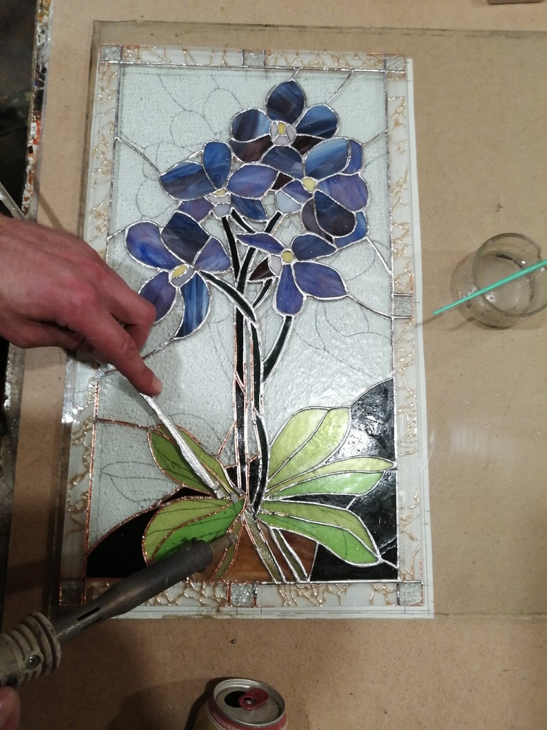 May include: A stained glass panel depicting a cluster of blue flowers with green leaves. The panel is being assembled with a soldering iron and solder.
