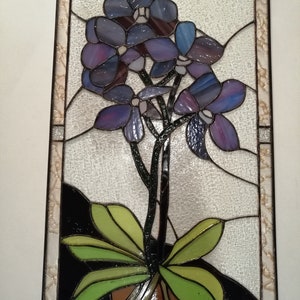May include: A stained glass window featuring a purple orchid with green leaves in a brown pot. The orchid is set against a white background with a black border.