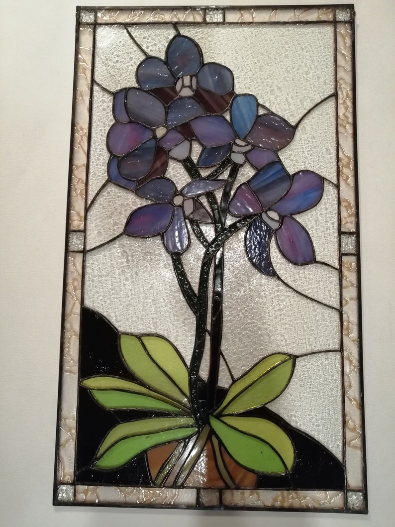 May include: A stained glass window featuring a purple orchid with green leaves in a brown pot. The orchid is set against a white background with a black border.