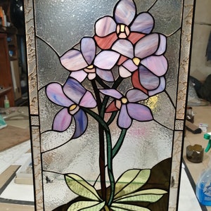 May include: A stained glass window featuring a bunch of purple orchids with green leaves and a brown pot. The orchids are clustered together with their petals overlapping.