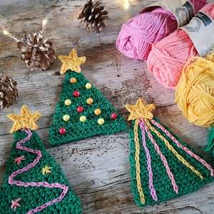 May include: Three crocheted Christmas tree ornaments in green with yellow stars and pink and yellow accents. The ornaments are on a wooden surface with yarn balls in pink, yellow, and white.