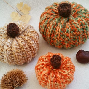 Puede incluir: Tres calabazas de ganchillo hechas a mano en tonos naranja, verde y beige, cada una con un tallo marrón. Las calabazas están dispuestas sobre una superficie blanca con follaje otoñal y una castaña.