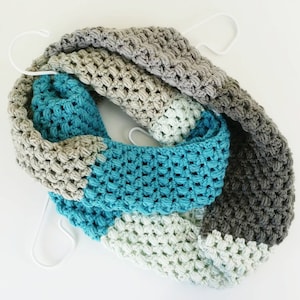 May include: A crocheted infinity scarf in shades of gray, teal, and white. The scarf is made with a textured stitch and has a chunky, cozy look.