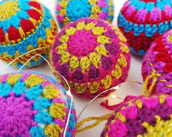 May include: A set of six crocheted ornaments in shades of pink, purple, blue, yellow, and red. The ornaments are decorated with a sparkly finish and feature a geometric pattern.