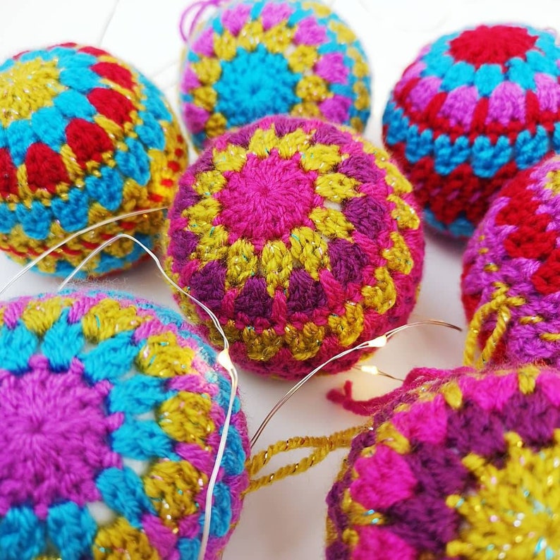 May include: A set of six crocheted ornaments in shades of pink, purple, blue, yellow, and red. The ornaments are decorated with a sparkly finish and feature a geometric pattern.