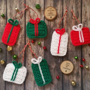 May include: Six crocheted gift box ornaments in red, green, and white with white ribbon bows. The ornaments are hanging from twine on a wooden background. The text "We wish you a Merry Christmas" and "With love at Christmas" are on two wooden circles.