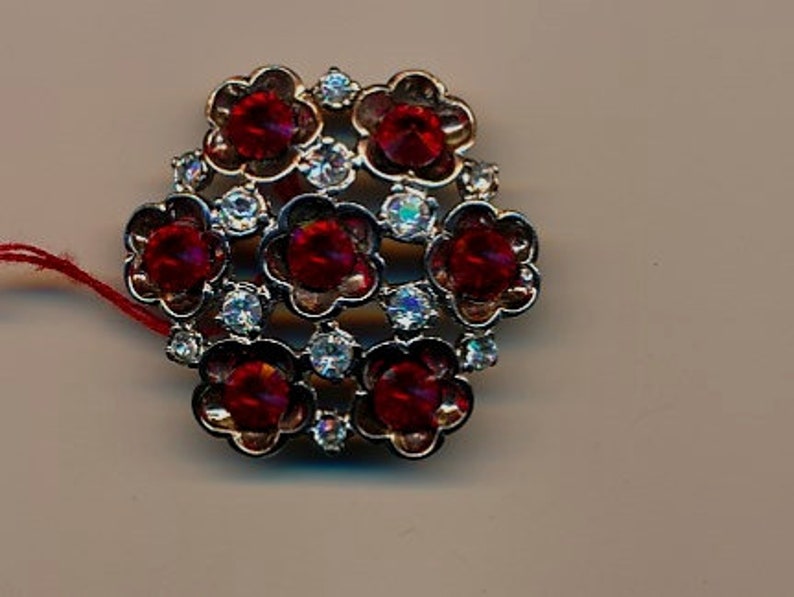 May include: A silver-toned brooch featuring a cluster of red and clear rhinestones in a flower design.