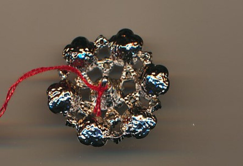 May include: A silver-toned metal embellishment with a red thread attached. The embellishment is shaped like a flower with a circular centre and six petals. Each petal has a raised, round bead at the end.