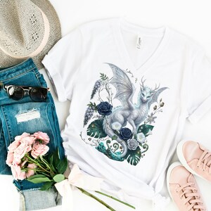 May include: A white V-neck t-shirt featuring a graphic of a silver dragon surrounded by dark blue roses and green tropical leaves. The outfit includes blue distressed denim jeans, a tan fedora hat, black sunglasses, pink sneakers, and pink roses.