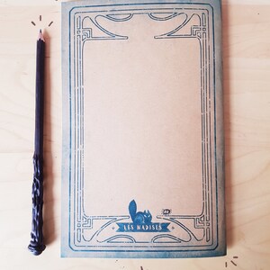 May include: A brown notebook with a blue decorative border and a small illustration of a cat and a bug. The notebook has the text "LES NADISES" printed on the bottom.