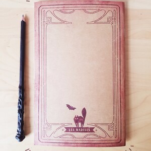 May include: A brown notebook with a decorative border and a black cat and bat illustration. The notebook has the text "LES NADISES" printed on the front.