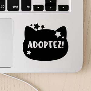 May include: Black and white sticker of a cat with stars and the text "ADOPTEZ!" on a white laptop.
