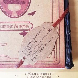 May include: A brown notebook with a witch illustration on the cover. The notebook is closed and has a brown paper tag attached to it. The tag says "1 Crayon Baguette 2 Carnets 2x48 pages lign&eacute;es - 21,6x13,95cm Papier 100% Recycle". The notebook is next to a black wand pencil.