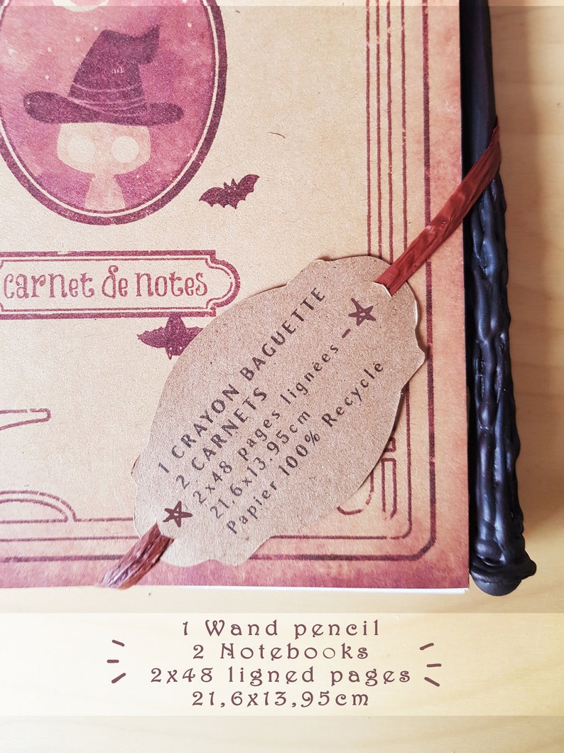 May include: A brown notebook with a witch illustration on the cover. The notebook is closed and has a brown paper tag attached to it. The tag says "1 Crayon Baguette 2 Carnets 2x48 pages lign&eacute;es - 21,6x13,95cm Papier 100% Recycle". The notebook is next to a black wand pencil.