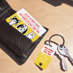 May include: A yellow and white card with cartoon pet illustrations is in a brown leather wallet. The card reads "MY PET IS HOME ALONE" and "MON ANIMAL EST SEUL CHEZ MOI." A matching keychain with the same text and a paw print is next to the keys.