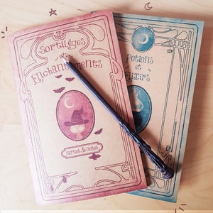 May include: Two vintage-style notebooks with decorative borders. The left notebook is red and says "Sortileges et Enchantements" with a witch's hat and bats. The right notebook is blue and says "Potions et Flixirs" with a potion bottle and a crescent moon. A black pencil is lying on top of the notebooks. The notebooks are made with 100% recycled paper and are lead-free.