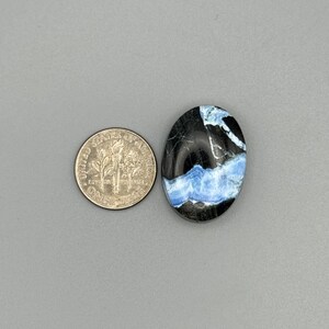 Könnte beinhalten: Ein ovaler Cabochon-Edelstein in Schwarz und Blau mit einem weißen und blauen Muster. Der Edelstein ist etwa so groß wie eine 10-Cent-Münze.
